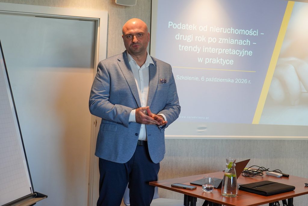 mężczyzna stoi, mówi, za nim wyświetla się slajd o podatkach od nieruchomości, szkolenie